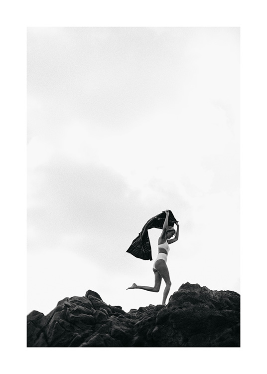  - Black and white photograph of a woman in a white bikini, running over rocks holding a towel over her head