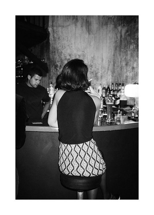  – Black and white photograph of a woman sitting at a stool at a bar, smoking a cigarette