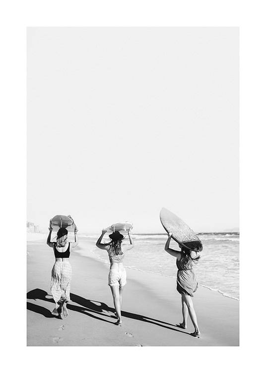  – Black and white photograph of three girls holding surfboards above their heads on the beach