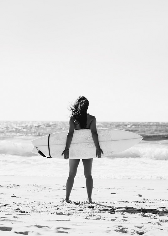  – Black and white photograph of a girl holding a surfboard behind her with the ocean in the background
