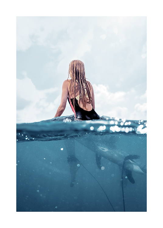  – Photograph of a woman sitting on a surfboard in the ocean, seen from behind