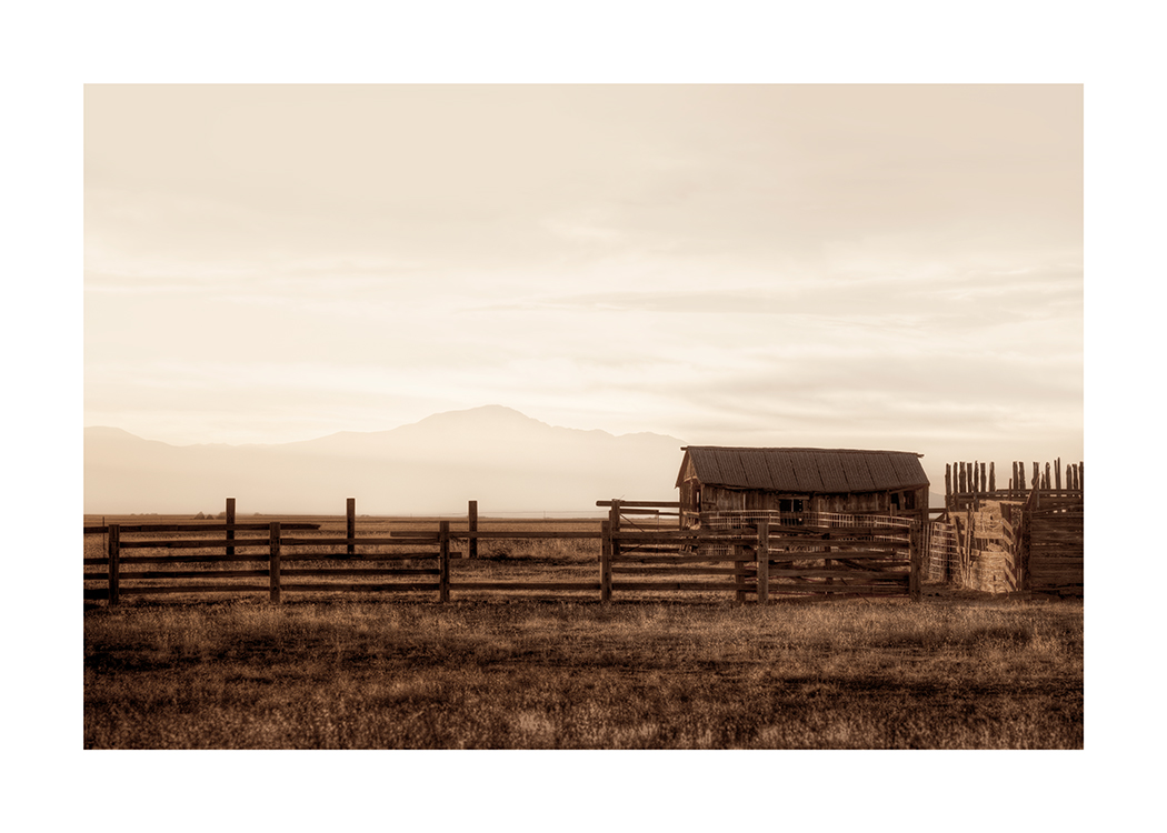 – A photograph of a barn and wooden fences in a brown field, with mountains and a light sky in the background