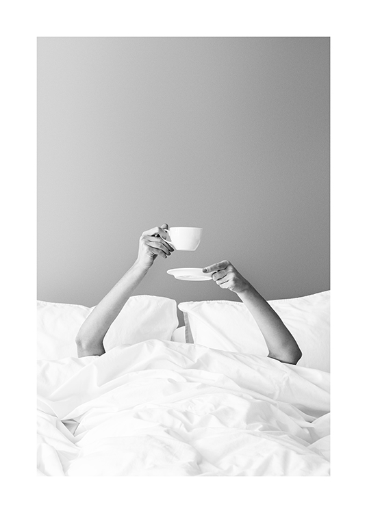 – Black and white photograph of a woman holding up a cup of coffee in bed from underneath the duvet cover